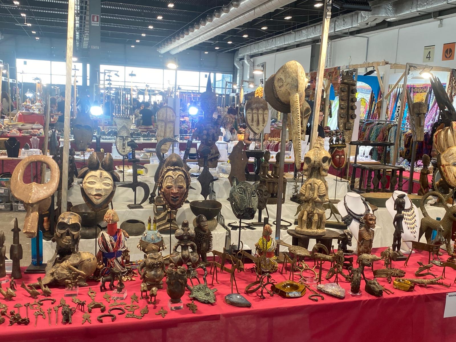 Banco espositivo in fiera con maschere, figure in legno e piccoli bronzi su un tavolo rosso